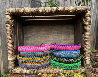 Puede incluir: Una colección de ocho collares de paracord trenzado dispuestos en dos pilas. Los collares presentan patrones vibrantes en rosa neón, morado, naranja, azul, verde, amarillo y negro, cada uno con un resistente anillo en D de metal negro.