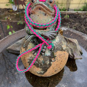 May include: A braided dog leash made of neon pink and dark green cord. The leash features a silver-toned metal swivel snap hook at one end and a looped handle at the other, showcasing a consistent woven pattern throughout the entire length.