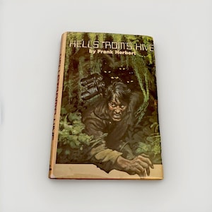 May include: A hardcover book titled Hellstrom's Hive by Frank Herbert. The cover features a dark, green-toned illustration of a person emerging from dense foliage with binoculars, while glowing eyes peer from the shadows in the background.