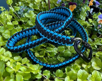 Peut inclure: Une laisse pour chien enroulée, fabriquée en paracorde noire et bleu vif avec un motif tressé. La laisse est dotée d'un mousqueton en métal noir à une extrémité, portant l'inscription 12kN, servant de point d'attache sécurisé.