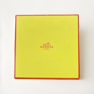 May include: A square, bright yellow box with a vibrant orange border. The center of the lid features the orange text HERMÈS PARIS accompanied by a small graphic illustration of a horse-drawn carriage, all printed in a clean, centered layout.