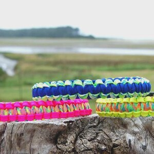 May include: Four colorful paracord bracelets rest on a weathered wooden surface. The bracelets feature vibrant hues of pink, blue, yellow, and green, with intricate woven patterns. The background shows a blurred landscape with water and greenery.