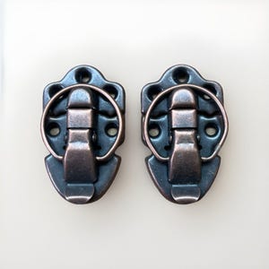 May include: Two antique bronze-colored metal latches. Each latch has a decorative shield-shaped base with multiple holes for mounting. A hinged clasp and a circular handle complete the design. Ideal for vintage boxes or craft projects.