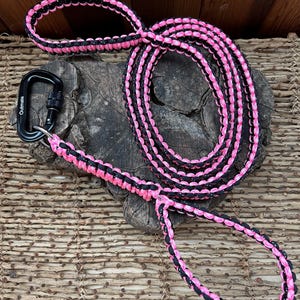 Op de afbeelding: Een roze en zwarte paracord hondenriem met een zwarte karabijnhaak. De riem is gemaakt van gevlochten koord en heeft een lus handvat. De karabijnhaak is bevestigd aan de riem met een veilig vergrendelingsmechanisme. De riem wordt getoond op een houten oppervlak.