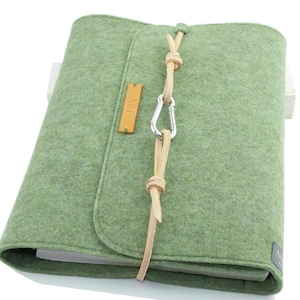 Könnte beinhalten: Eine hellgrüne Filz-Notizbuchhülle mit Lederriemenverschluss und silbernem Karabiner. Die Hülle hat ein kleines, rechteckiges Lederetikett und ein aufgesticktes Logo in der unteren rechten Ecke. Das Notizbuch ist geschlossen.