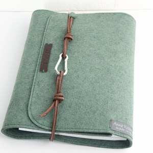 May include: A sage green felt journal cover with a leather strap closure and a silver carabiner. The cover has a rounded flap and a small leather tag. The journal is closed and resting on a white surface.