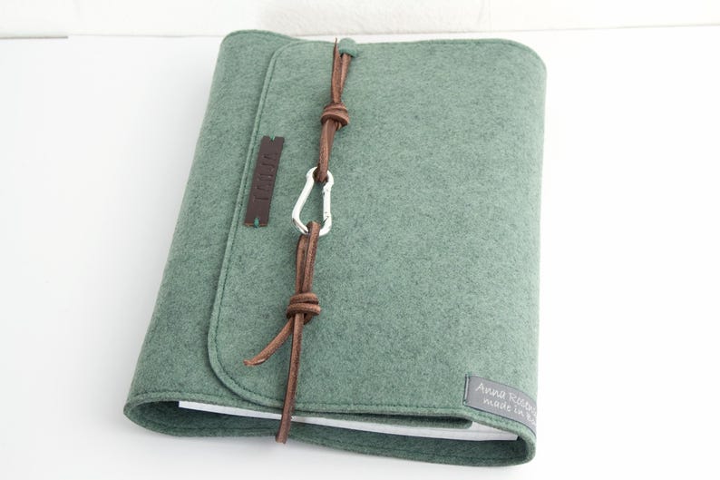 May include: A sage green felt journal cover with a leather strap closure and a silver carabiner. The cover has a rounded flap and a small leather tag. The journal is closed and resting on a white surface.
