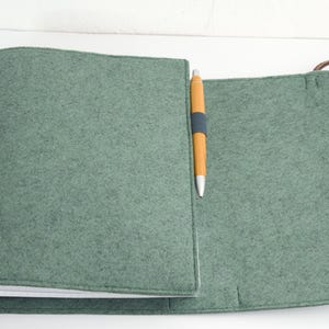 May include: A sage green felt notebook cover with a matching pen. The cover is open, revealing the interior pages. A brown leather strap is attached to the cover. The pen is yellow with a blue band.