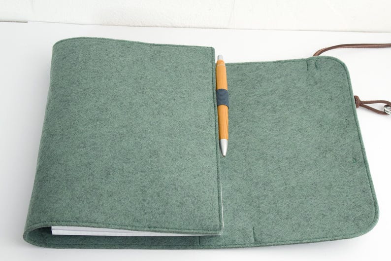 May include: A sage green felt notebook cover with a matching pen. The cover is open, revealing the interior pages. A brown leather strap is attached to the cover. The pen is yellow with a blue band.