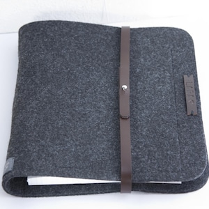 Könnte beinhalten: Ein graues Filz-Portfolio mit einem braunen Lederriemen und einer silbernen Schnalle. Das Portfolio hat ein Etikett an der Seite, auf dem "JUNK" steht.