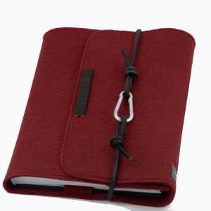 Könnte beinhalten: Ein bordeauxfarbener Notizbuchumschlag aus Filz mit einem schwarzen Lederriemen und einem silbernen Karabinerhaken. Der Umschlag hat ein kleines Leder-Etikett mit dem eingeprägten Wort "NOTES".