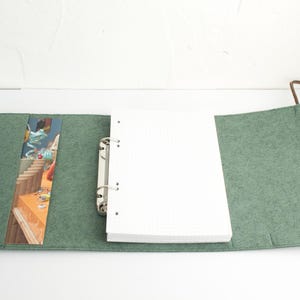 May include: Open green felt journal with a white grid paper notebook and a silver binder. A colorful bookmark is visible inside the journal. A gold and black pen rests on the left side of the journal.