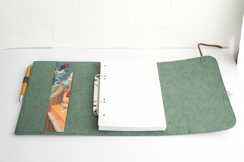 May include: Open green felt journal with a white grid paper notebook and a silver binder. A colorful bookmark is visible inside the journal. A gold and black pen rests on the left side of the journal.