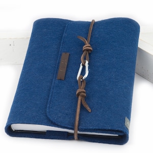 Könnte beinhalten: Ein blauer Filz-Notizbuchumschlag mit einem braunen Lederriemen und einem silbernen Verschluss. Der Umschlag hat ein kleines braunes Lederetikett mit dem eingeprägten Wort "Felt".