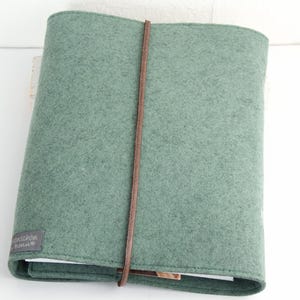 May include: A sage green felt journal cover with a brown leather strap closure. The cover has a rectangular shape and a small gray label with white text on the bottom left corner. The journal is closed.