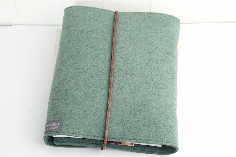 May include: A sage green felt journal cover with a brown leather strap closure. The cover has a rectangular shape and a small gray label with white text on the bottom left corner. The journal is closed.