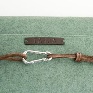 May include: Close-up of a sage green felt bag with a brown leather strap and silver carabiner clasp. The bag has a small brown leather tag with the name "TANJA" embossed on it. The bag is made of a soft, textured material.
