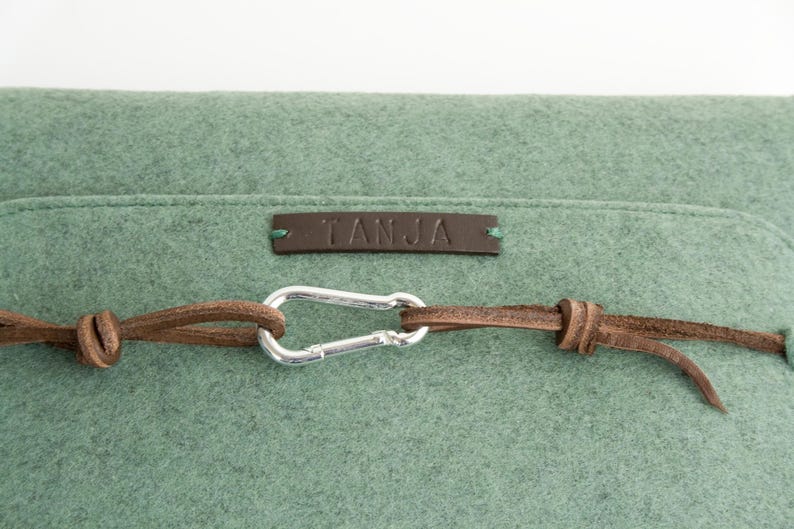May include: Close-up of a sage green felt bag with a brown leather strap and silver carabiner clasp. The bag has a small brown leather tag with the name "TANJA" embossed on it. The bag is made of a soft, textured material.