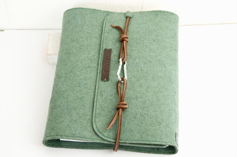 May include: A sage green felt journal cover with a leather closure. The cover is secured with a brown leather cord and a silver carabiner. A small brown leather tag is attached to the front. The journal cover is closed.