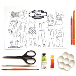 May include: A black and white printable paper doll set with the name "Adele" and the text "Paper Doll Par Carambouilles". The set includes a doll and various outfits to color and cut out. The image also shows scissors, paintbrushes, paint, and a palette.
