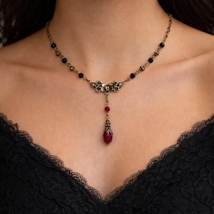 May include: A vintage-style necklace with a bronze-toned chain and floral pendant. The necklace features black beads and a dangling red teardrop-shaped pendant. The intricate design adds a touch of elegance and gothic charm.