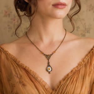 May include: A bronze-toned metal necklace featuring a delicate chain with small amber-colored beads. The pendant is an elongated teardrop shape with an ornate metal frame surrounding a central white oval inlay displaying a small yellow floral design.