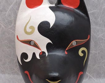 May include: A fox mask featuring a split black and white design. The mask has pointed ears with red inner accents, almond-shaped eye openings with red and gold outlines, and decorative gold swirls on the cheeks. A thin red line forms a smile.