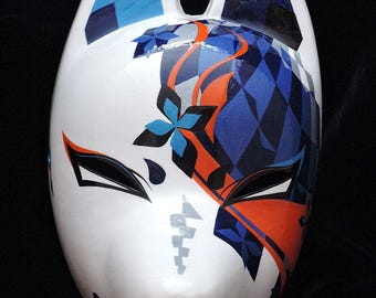 May include: A white fox mask with a stylized design. The mask features blue and orange geometric patterns, including diamonds and floral accents. The ears are painted with blue and black checkered patterns. Black eye details and a curved mouth complete the design.