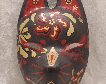 May include: A fox mask featuring a dark charcoal base with intricate hand-painted floral patterns in red and gold. The design includes pointed ears, almond-shaped eyes, a central nose detail, and stylized whiskers, all rendered in a symmetrical layout.