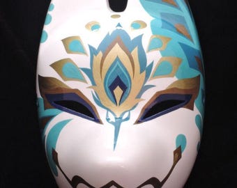 Puede incluir: Una máscara de zorro blanca con detalles pintados en tonos turquesa, azul oscuro y dorado. El diseño incluye patrones simétricos alrededor de los ojos, un motivo floral central en la frente y una línea de boca negra en zigzag.