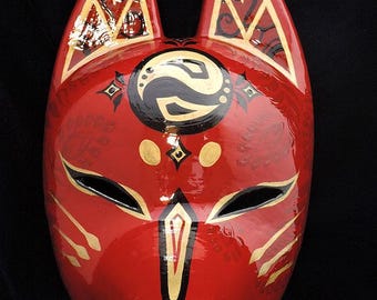 Puede incluir: Una máscara de zorro roja brillante con orejas puntiagudas que presentan patrones geométricos dorados y negros. El rostro muestra un símbolo circular central en negro y oro, aberturas para los ojos y marcas de bigotes doradas en las mejillas.