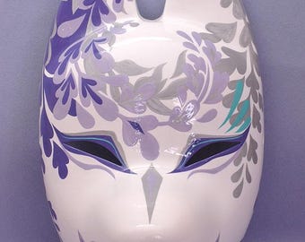Puede incluir: Una máscara de cerámica blanca con un diseño de zorro estilizado. La máscara presenta patrones florales morados y grises, con aberturas para los ojos negras y un diseño de boca en zigzag. Las orejas son puntiagudas y decoradas con patrones florales morados.