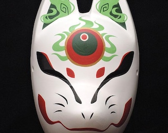 May include: A white fox mask featuring pointed ears, black almond-shaped eye cutouts, and red markings on the cheeks and forehead. A central circular design in dark green and red sits on the forehead, surrounded by green flame-like patterns.