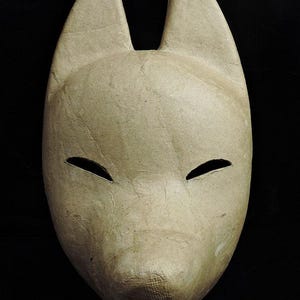 Peut inclure: Un masque de renard beige avec une surface lisse et texturée, des oreilles pointues et des ouvertures pour les yeux étroites en forme d'amande. Le masque présente un museau arrondi et un design simple, avec une finition semblable à du papier.