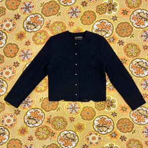 May include: A dark navy blue cardigan with a round neckline and long sleeves. The button-down sweater has small, white buttons and a tailored fit. The garment is displayed on a patterned background with orange and yellow floral designs.