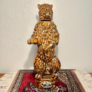 Puede incluir: Un decantador de cerámica con forma de oso de pie con un acabado marrón brillante. La cabeza del oso sirve como tapa extraíble. Una etiqueta en la base dice Ezra Brooks 12 Years Old 90 Proof Real Sippin Whiskey. La figura presenta detalles de pelaje texturizado.