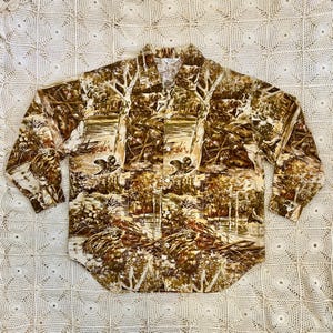 May include: Button-down shirt with a nature-themed print in shades of brown, beige, and green. The long-sleeved shirt features a detailed landscape design with trees, water, and wildlife. The shirt is laid flat on a white, textured surface.