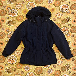 May include: A navy blue hooded jacket with a drawstring waist and a zipper closure. The jacket has a hood and a small logo on the upper left sleeve. The jacket is on a floral patterned background.