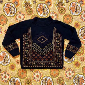 Könnte beinhalten: Ein schwarzer Pullover mit hohem Kragen und langen Ärmeln. Die Vorderseite des Pullovers zeigt ein geometrisches Design in Braun- und Goldtönen. Die Ärmel haben ebenfalls ein ähnliches Muster in der Nähe der Manschetten. Der Pullover wird auf einem floral gemusterten Hintergrund präsentiert.