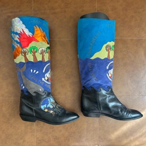 May include: A pair of knee-high black leather boots featuring colorful embroidered panels. The shafts display scenes of forests, flames, and a whale with text reading STOP POLLUTION and SAVE THE WHALES. The foot area includes a circular graphic.