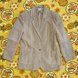 May include: A light brown suede blazer with a notched collar, two front pockets, and two buttons. The blazer has long sleeves and a tailored fit. The jacket is a neutral color and suitable for various outfits.