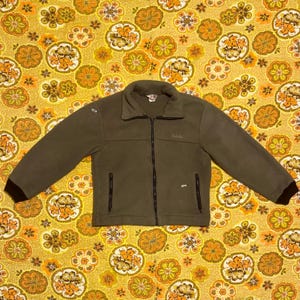 Puede incluir: Chaqueta polar verde oliva con cremallera negra de largo completo y dos bolsillos laterales. La chaqueta tiene cuello y puños marrones oscuros. El nombre de la marca "Cabela's" está bordado en el pecho.
