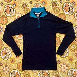 May include: A black long-sleeved shirt with a teal collar and zip. The shirt features a half-zip closure and a teal-coloured interior collar. The shirt is made of a soft, comfortable material.