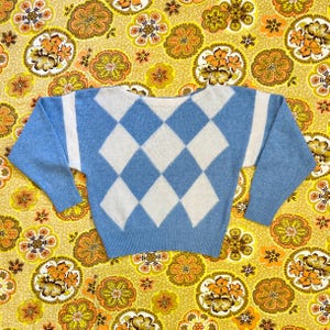 May include: A light blue sweater featuring a white diamond pattern. The sweater has long sleeves and a crew neck. The background is a yellow floral pattern.