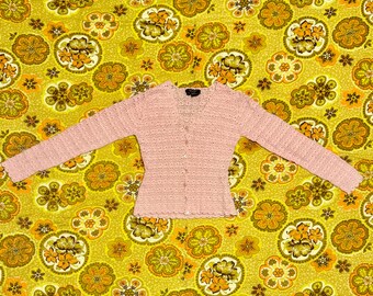 Può includere: Un cardigan rosa chiaro a maniche lunghe con chiusura a bottoni. Il maglione presenta un delicato design a maglia aperta e un orlo smerlato. Lo sfondo è un motivo floreale giallo.