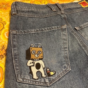 Pode incluir: Jeans azuis com um patch decorativo no bolso de trás. O patch apresenta um urso de desenho animado usando um saco de papel na cabeça, segurando uma garrafa. O urso é branco com uma mancha preta.