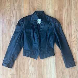May include: A black leather cropped jacket with a single button closure and a small label that reads "Willowbrook Furs" on the front.