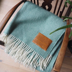 May include: A teal green herringbone throw blanket with a white fringe. The blanket is draped over a wooden chair with a leather label attached to it. The label has text on it.