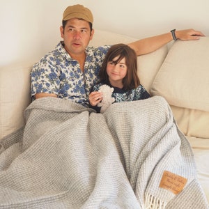 May include: A light grey waffle-weave blanket with a fringed edge, draped over a sofa. A leather tag on the blanket reads "Daddy & Sully's Movie night Snuggle blanket". A man and a child are seated on the sofa.