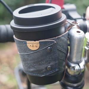 Op de afbeelding: Een zwarte reiskoffiemok met deksel in een donkerblauwe leren fietsbekerhouder. De houder is bevestigd aan het stuur en heeft een klein, rond, bruin label met het woord "Stapo".